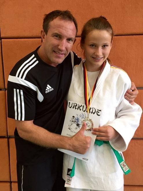 Mann in Sportkleidung umarmt lächelnd ein Mädchen in Judokleidung, das eine Urkunde und Medaille hält.