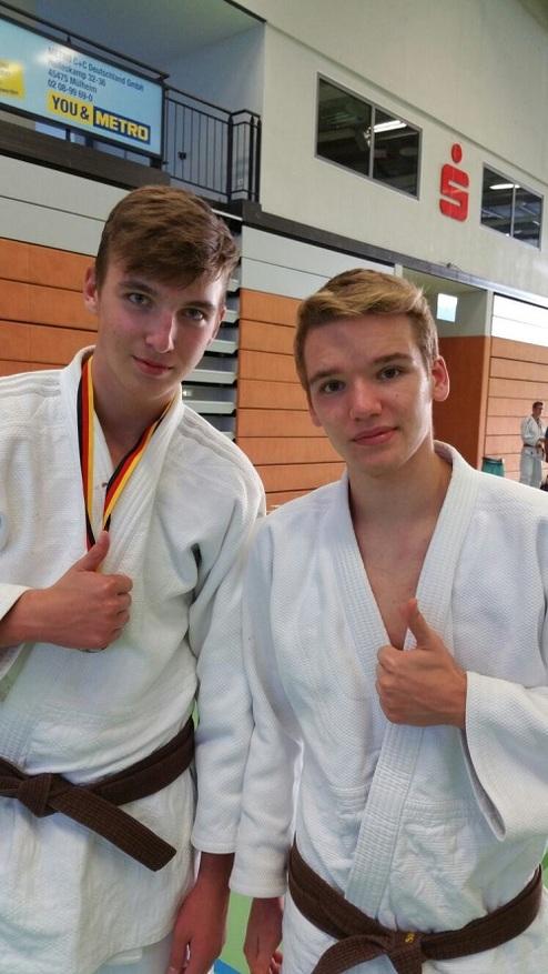 Zwei junge Judoka in weißen Judogi zeigen Daumen hoch, eine Medaille hängt am Hals des Linksstehenden.