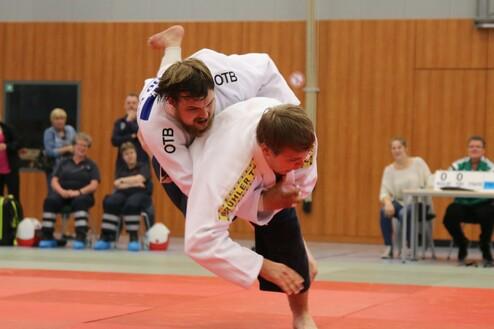 Zwei Judokas in Wettkampfhaltung auf einer roten Wettkampfimprovisation, Zuschauer im Hintergrund.
