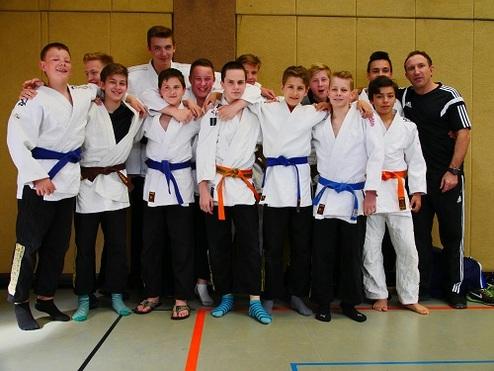 Gruppe von zwölf Judoka in weißen Gi mit blauen und orangefarbenen Gürteln, posierend in einer Sporthalle.