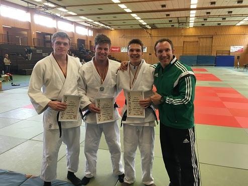 Vier Judo-Kämpfer in Traininganzügen stehen auf einer Mattenfläche und halten mit Stolz ihre Urkunden in den Händen.