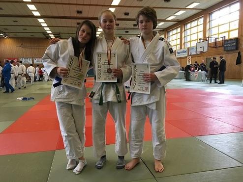 Drei Judoka in weißen Anzügen halten Urkunden, im Hintergrund eine Sporthalle mit weiteren Teilnehmern auf Matten.