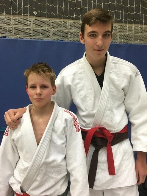 Zwei Jungen in Judoanzügen stehen vor einer blauen Wand; der Ältere trägt einen roten Gürtel, der Jüngere einen weißen.