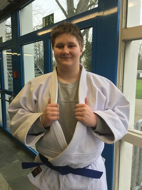 Junger Mensch in Weißem Judoanzug mit blauer Gürtelfarbe, steht in einer Sporthalle und lächelt.