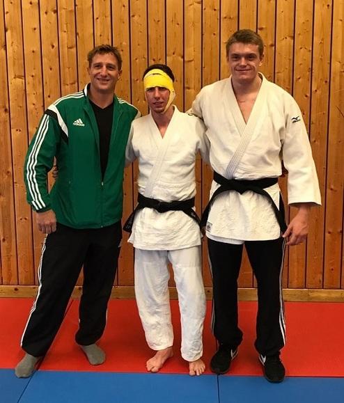 Drei Männer stehen in Judogewändern auf einer Mattenfläche, Holzwand im Hintergrund. Zwei tragen schwarze Gürtel, einer gelb.