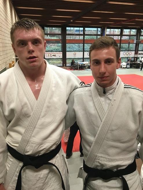 Zwei Judoka in weißen Gi mit schwarzen Gürteln stehen nebeneinander in einer Sporthalle auf einer Judomatte.