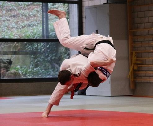 Zwei Judoka in Weiß und Schwarz führen einen Wurf im Training auf einer roten Matte in einem Dojo aus.