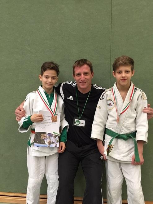Zwei Jungen in Judogis halten Medaillen und Urkunden, während ein Trainer in der Mitte lächelt. Hintergrund ist eine grüne Wand.