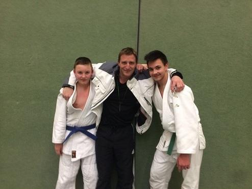 Drei Personen in Judogi stehen lächelnd zusammen auf einer grünen Matte, umarmt von einem Trainer in Sportkleidung.