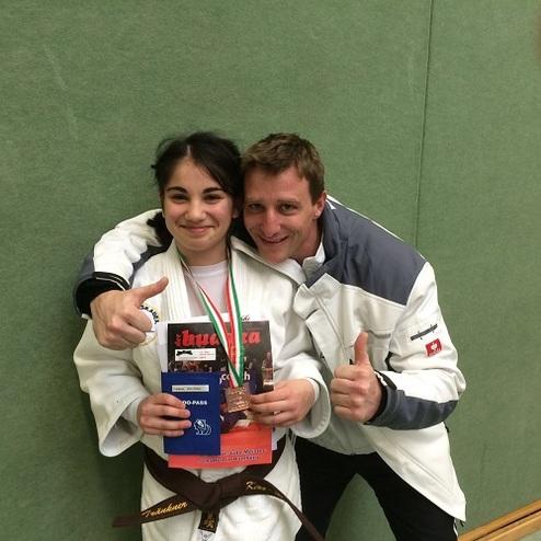 Ein junger Mädchen in Karateanzug hält Medaillen und Urkunden, während sie mit einem Trainer lächelt und Daumen hoch zeigt.