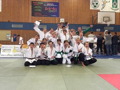 Gruppe von Jugendlichen in Judogis feiert mit Medaillen und Urkunden nach einem Wettkampf auf einer Judomatte.