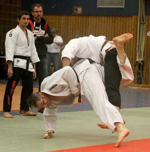 Zwei Judokas im Wettkampf; einer wirft den anderen auf eine Tatami, während Zuschauer im Hintergrund beobachten.