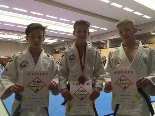 Drei Jugendliche in Judoanzügen holding Urkunden nach einem Wettkampf in einer Sporthalle.