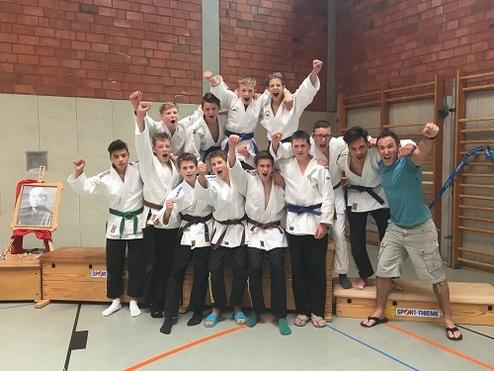 Gruppe von Jugendlichen in Judogi, posierend auf einer Matte, feiert jubelnd nach einem Wettkampf.