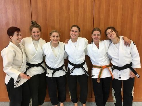 Sechs Frauen in Judoanzügen und schwarzen Gürteln stehen lächelnd vor einer Holzwand.
