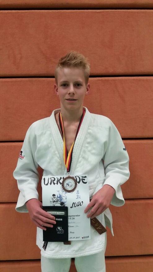 Junge im Judoanzug hält eine Urkunde und Medaille vor oranger Wand. Er trägt einen Teilnehmerausweis um den Hals.