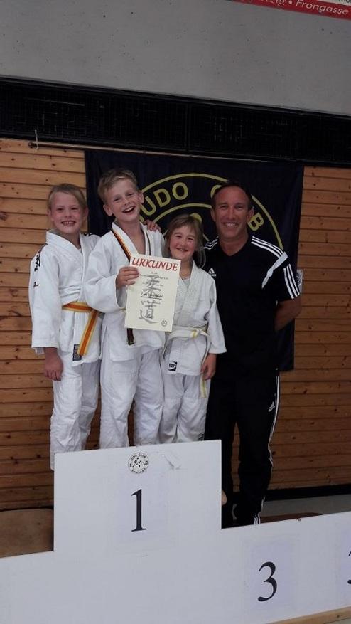 Kinder in Judoanzügen posieren mit einer Urkunde, während ein Trainer neben ihnen steht, auf einem Podium in einer Sporthalle.