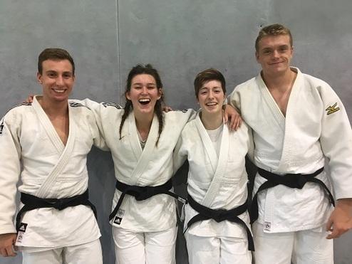 Vier Judoka in weißen Judogis mit schwarzen Gürteln stehen lächelnd vor einer grauen Wand.