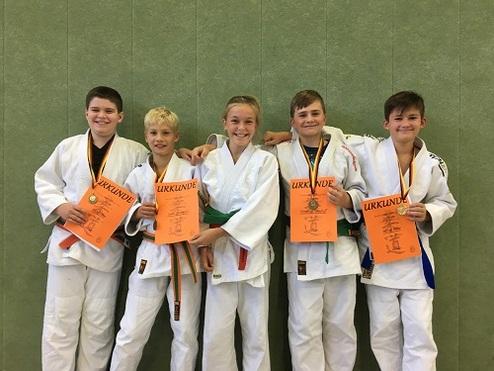 Fünf Kinder in Judogewändern halten stolz Urkunden und Medaillen auf einer glatten Wand hinter ihnen.