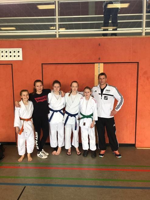 Gruppe junger Judoka in Trainingsanzügen posiert mit Trainer vor einer orangefarbenen Wand in einer Sporthalle.
