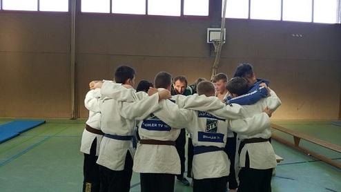 Dutzende von Jugendlichen in Judokleidung stehen im Kreis und umarmen sich in einer Sporthalle.