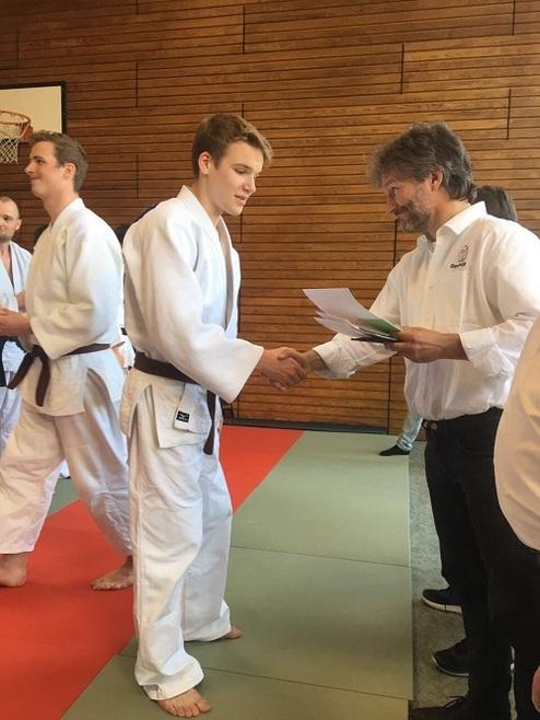 Junger Judoka in Weiß empfangen von einem Prüfer, während er eine Urkunde erhält. Karateanzüge und Sporthalle im Hintergrund.