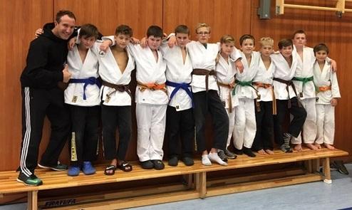 Gruppe von Jugendlichen in Judogi, stehend auf einer Bank, posiert mit einem Trainer in einem Sportraum.