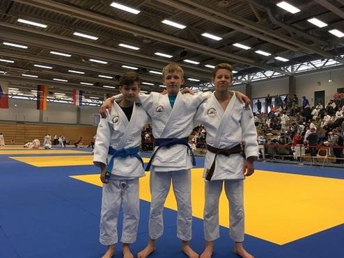 Drei jugendliche Judoka in weißen Gis mit blauer und brauner Gürtelstellung stehen auf einer Wettkampffläche.