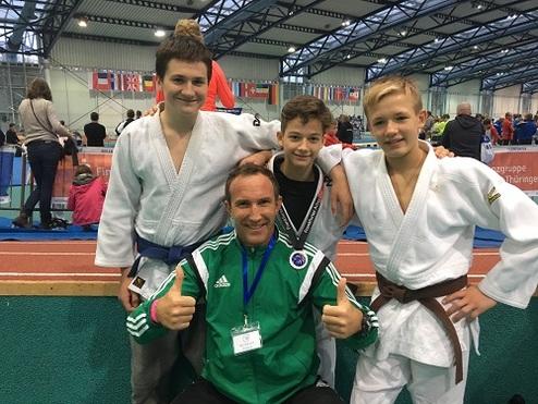 Drei Jungen in Judogis posieren mit einem Trainer und zeigen Daumen hoch, im Hintergrund ein Wettkampfszenario.