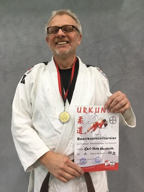 Mann in Judogi mit Medaille lächelt und hält eine Urkunde vom Bezirks-Einzelturnier in der anderen Hand.