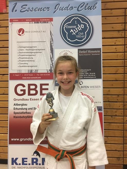 Mädchen in Judogi mit Medaille lächelt vor einem Plakat des Essener Judo-Clubs.