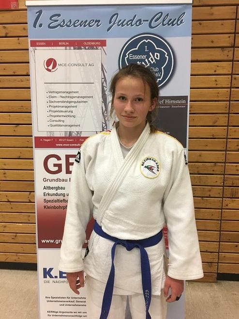 Mädchen im Judogi mit blauer Gürtelfarbe steht vor einem Banner des Essener Judo-Clubs.