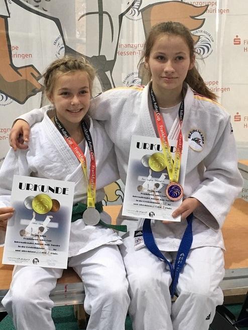 Zwei junge Judoka in Anzügen sitzen nebeneinander und halten Urkunden sowie Medaillen in der Hand.