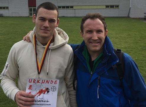 Zwei Männer stehen lächelnd im Freien, einer hält eine Urkunde, der andere trägt eine blaue Jacke.