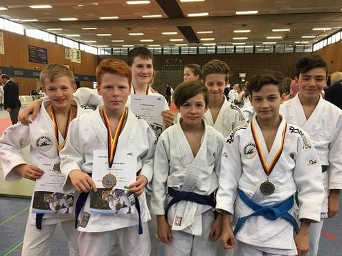 Sechs Kinder in Judokas, lächeln und halten Medaillen sowie Urkunden in einer Sporthalle.