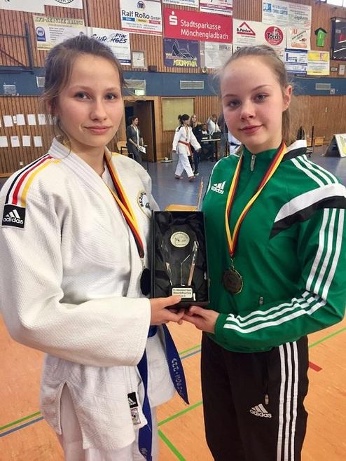 Zwei junge Judokas halten zusammen einen Pokal, während sie medailliert in einem Sportzentrum stehen.