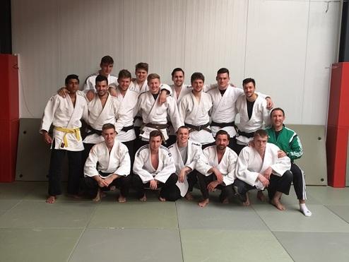 Gruppe von 14 Judokas in weißen Gi mit schwarzen und gelben Gürteln posieren auf einer Judomatte.