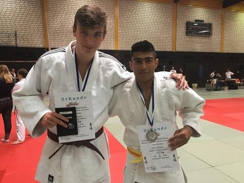 Zwei Judokas in weißen Gi halten ihre Urkunden und lächeln auf einer Wettkampfmatte. Hintergrund mit Zuschauern und Matten.