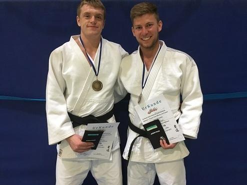 Zwei Judo-Sportler in weißen Judogi zeigen stolz ihre Urkunden und Medaille vor blauer Wand.