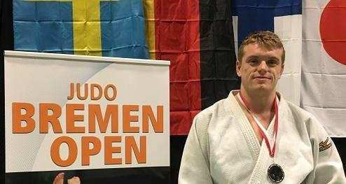 Junger Judoka in weißem Gi steht mit Medaille vor einem Schild „JUDO BREMEN OPEN“ und mehreren Fahnen im Hintergrund.