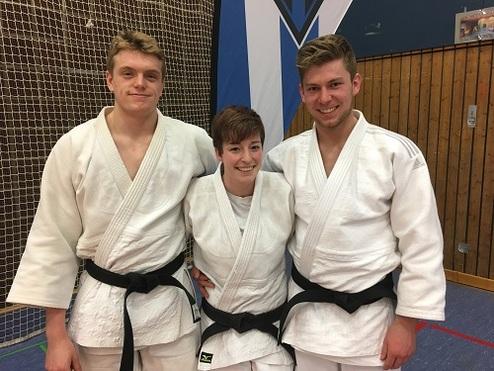 Drei Judoka in weißen Judogis mit schwarzen Gürteln posieren lächelnd vor einer Sporthalle.