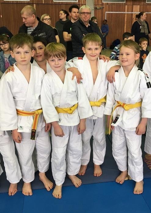 Fünf Kinder in weißen Gi mit gelben Gürteln stehen lächelnd auf einer Judomatte.