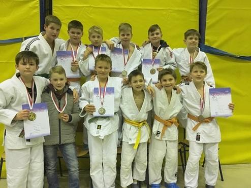 Gruppe von zwölf jungen Judokas in weißen Gi, mit Medaillen und Urkunden, vor gelber Wand.