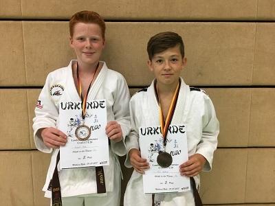 Zwei Jungen in Judokleidung halten Urkunden mit Medaillen, stehen vor einer hellen Wand.