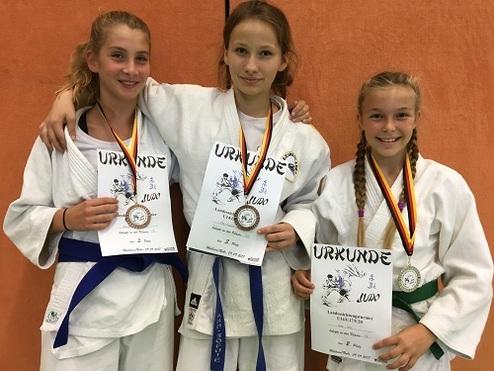 Drei Mädchen in Judogi halten Urkunden und Medaillen, stehen vor einer orangefarbenen Wand.