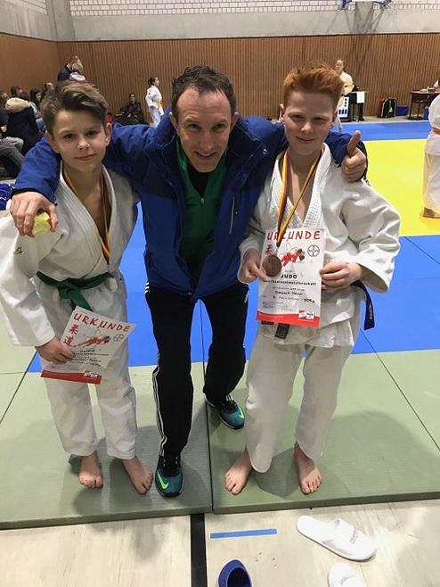 Zwei Jungen in Judogis halten Urkunden, während ein Trainer zwischen ihnen steht und beide umarmt.