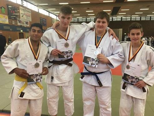Vier Judoka in weißen Judogis zeigen stolz ihre Medaillen und Urkunden nach einem Wettkampf in einer Sporthalle.