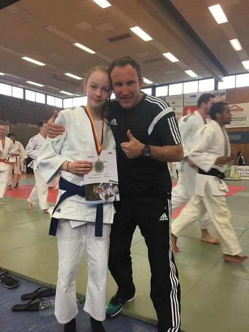 Mädchen in Judo-Gi mit Medaille und Urkunde, umarmt von Trainer in Sportkleidung, Judo-Training im Hintergrund.