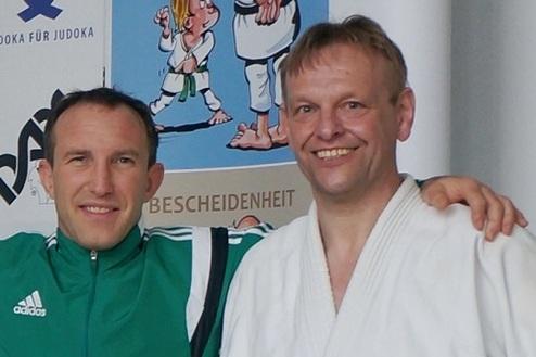 Zwei Männer lächeln in Sportbekleidung, einer trägt einen weißen Judo-Anzug, vor einem plakativ gestalteten Hintergrund.