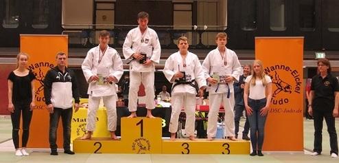 Judo-Siegerehrung mit drei Athleten auf dem Podium und Trainern an den Seiten, orangefarbene Banner im Hintergrund.